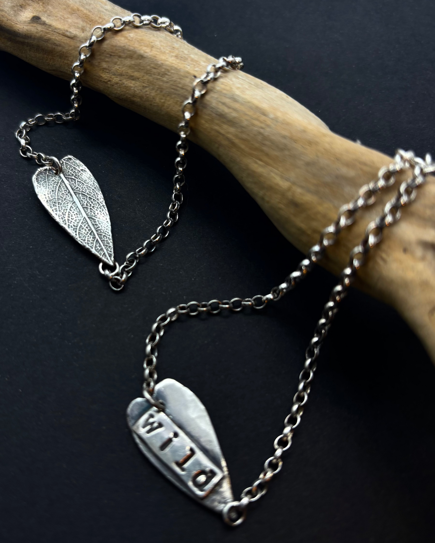 Reversible recycled silver bracelet with heart-shaped sage leaf textured charm and “Wild” engraving on the back, against a dark background with a branch. 