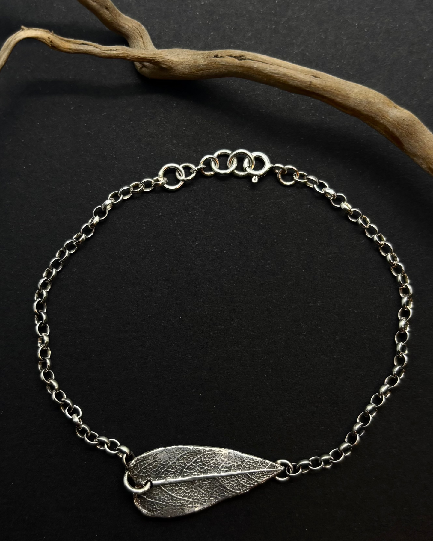 Reversible recycled silver bracelet with heart-shaped sage leaf textured charm and “Wild” engraving on the back, against a dark background with a branch. 