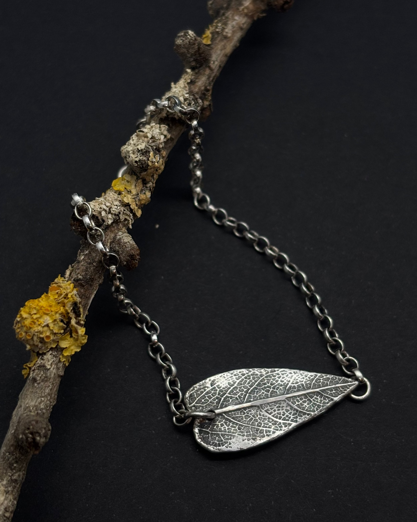 Reversible recycled silver bracelet with heart-shaped sage leaf textured charm and “Wild” engraving on the back, against a dark background with a branch. 