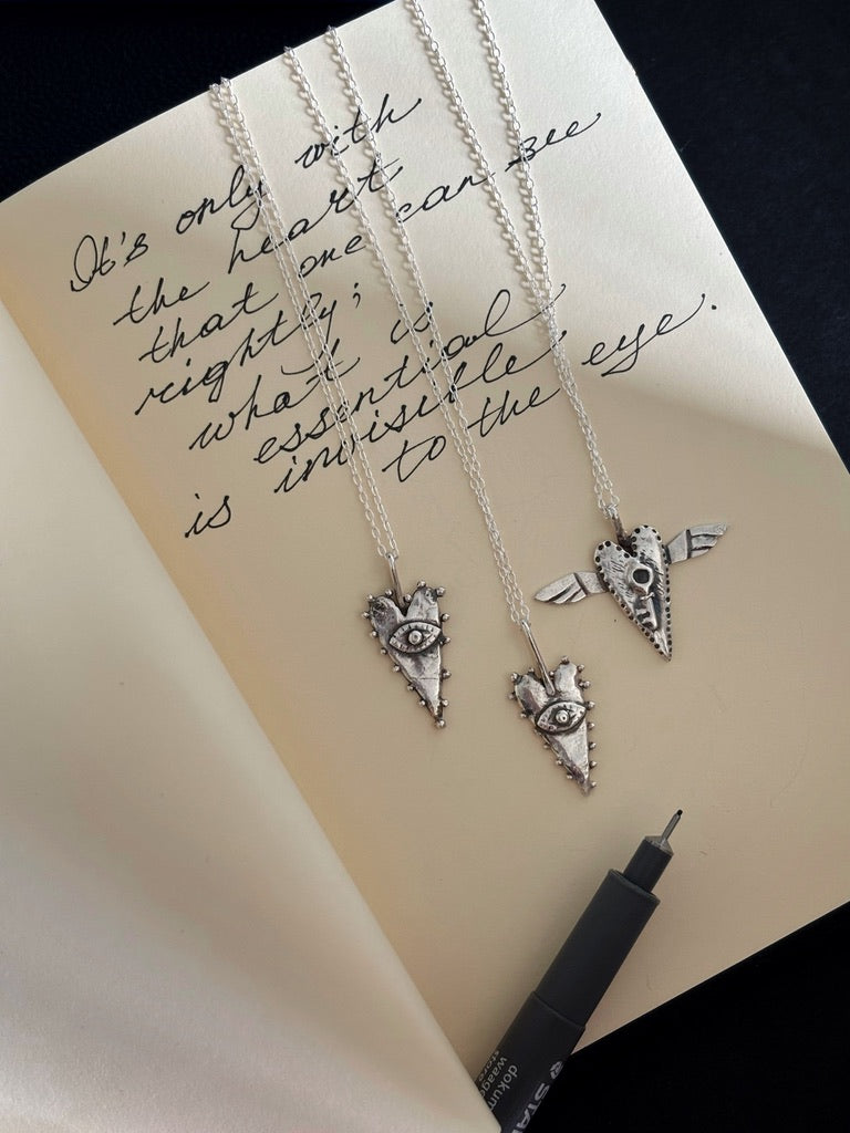 The All-Seeing Heart mystical silver pendant photographed on a notebook with the handwritten Little Prince quote, “It is only with the heart that one can see rightly; what is essential is invisible to the eye.” Tarot-inspired symbolic jewellery featuring an all-seeing eye and heart motif, inspired by magic, alchemy, intuition and hidden wisdom. Artistic silver talisman necklace with a whimsical, slightly naive aesthetic.