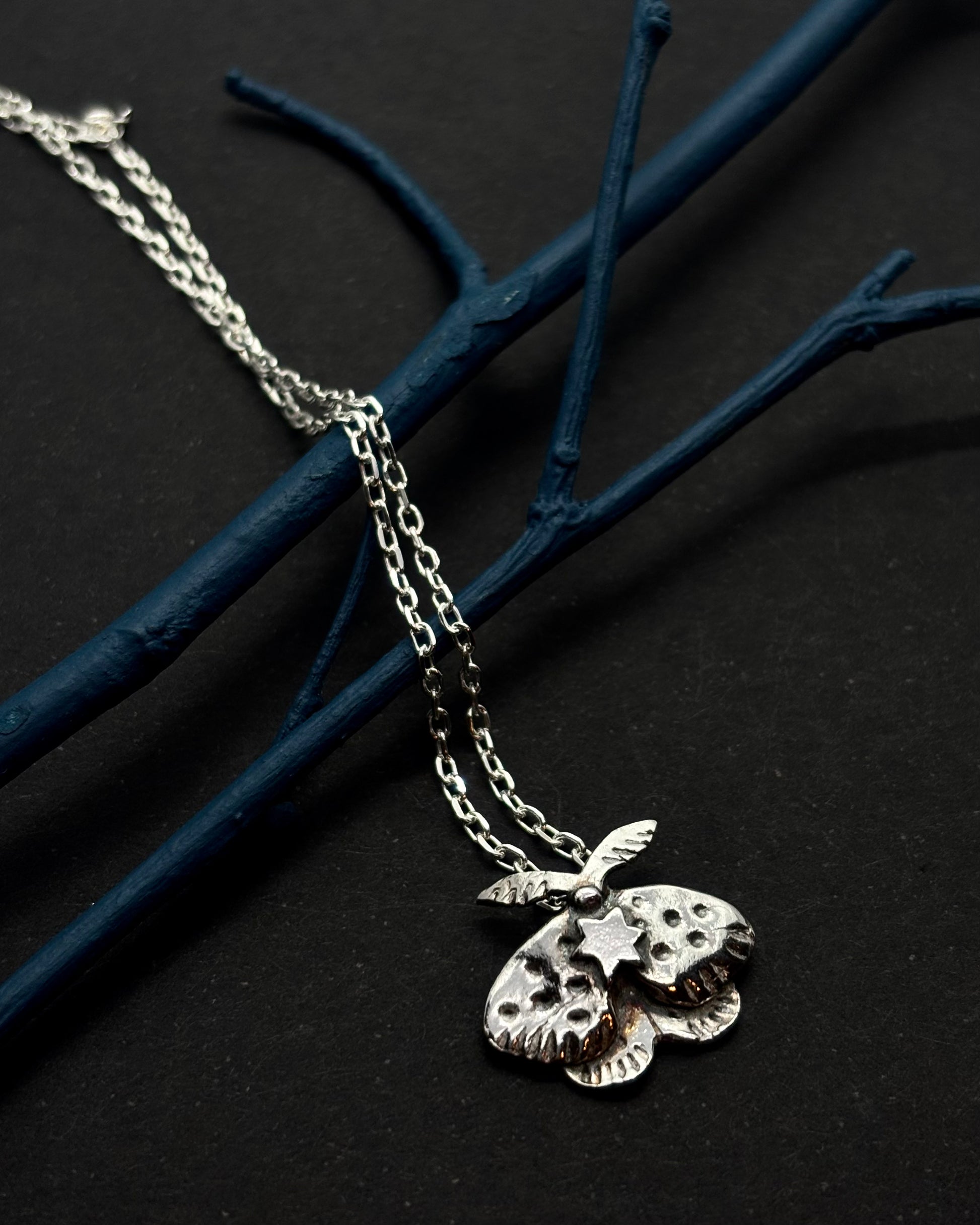 Handcrafted silver moth pendant with detailed wings, inspired by nocturnal nature and made in Ireland; on black background with twigs.