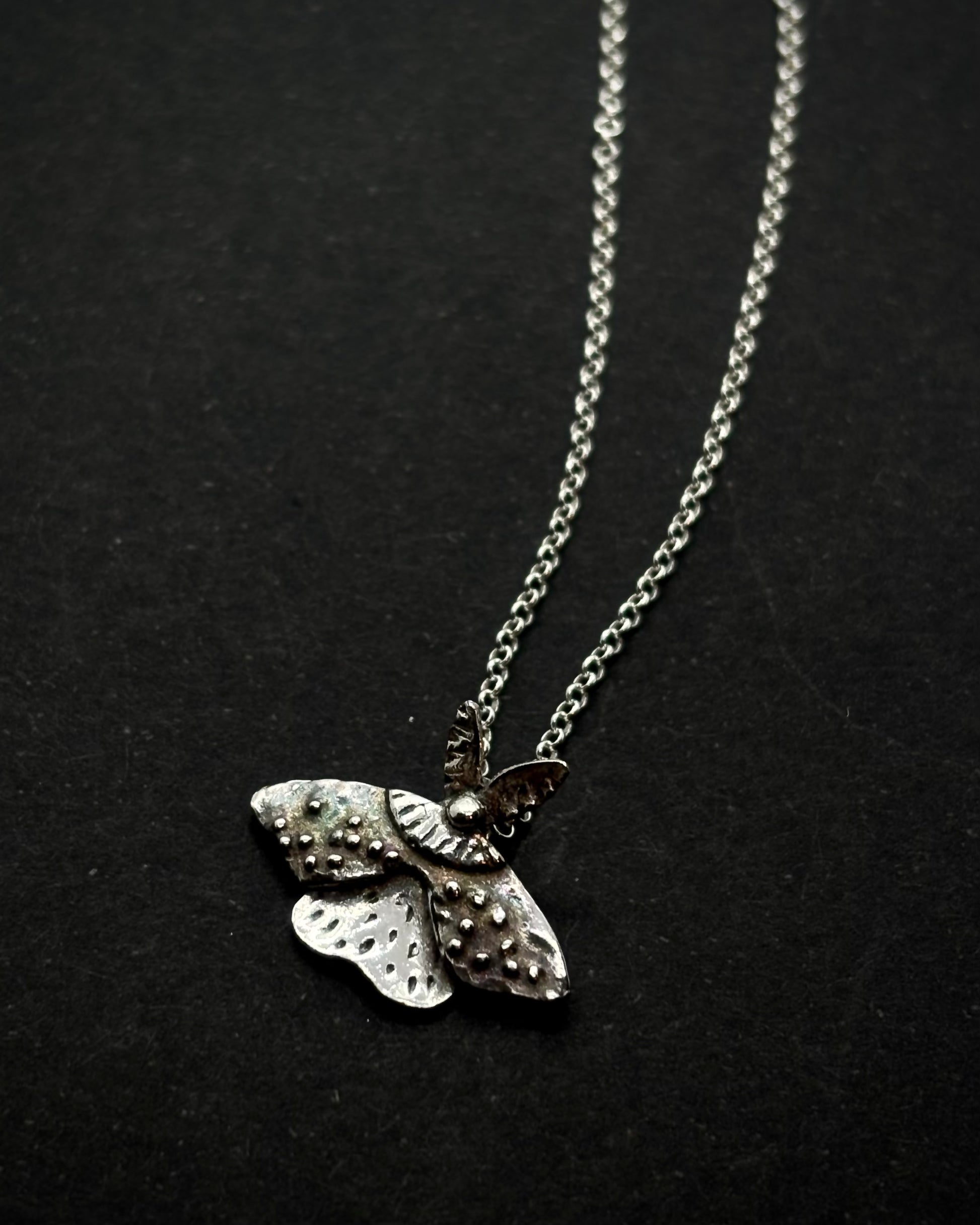 Handcrafted silver moth pendant with detailed wings, inspired by nocturnal nature and made in Ireland; on a chain, shon on dark background