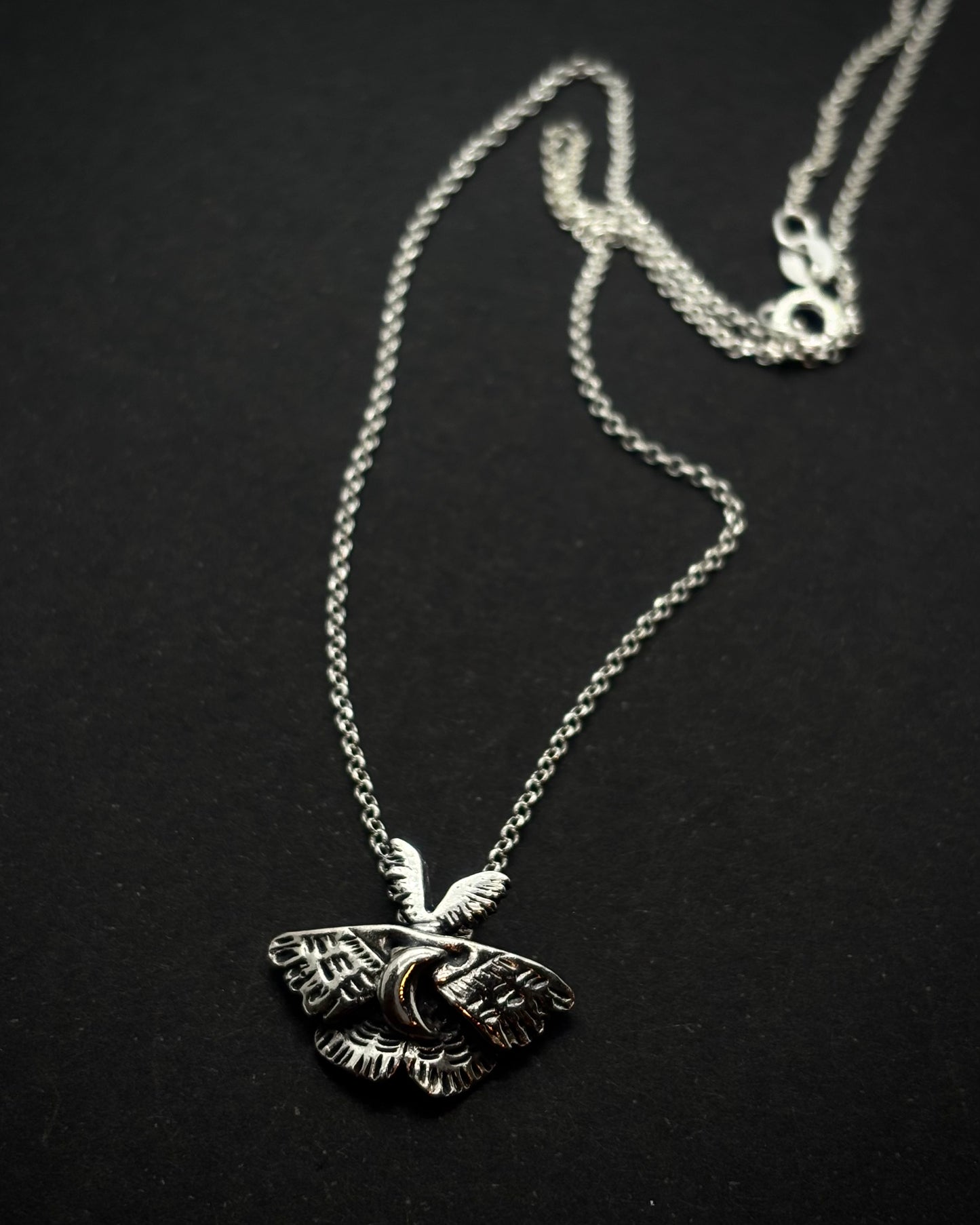 Handcrafted silver moth pendant with detailed wings, inspired by nocturnal nature and made in Ireland, shown on a chain, laid out on a black background