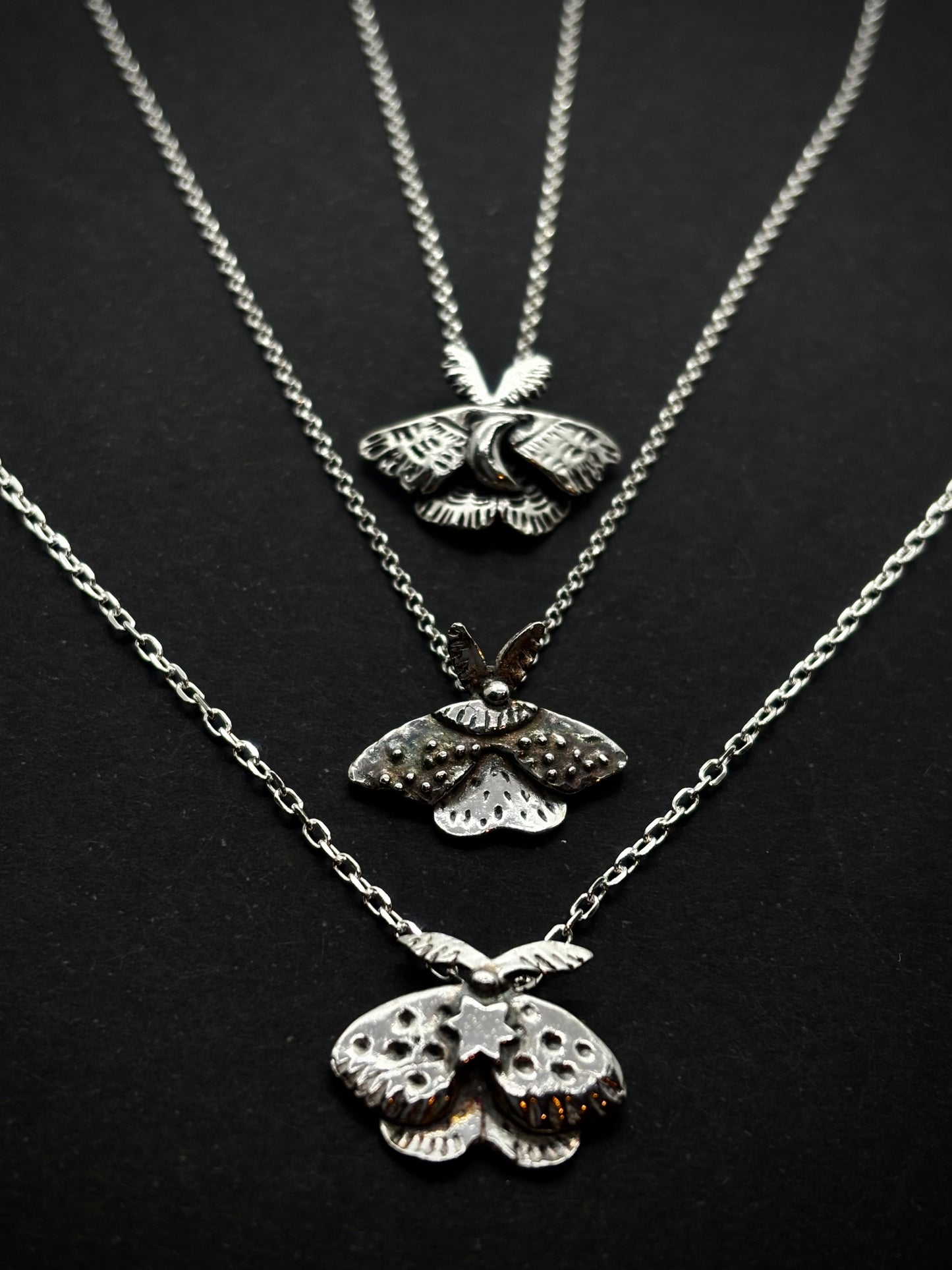 Handcrafted silver moth pendants with detailed wings, inspired by nocturnal nature and made in Ireland, on silver chains, laid out on black background.